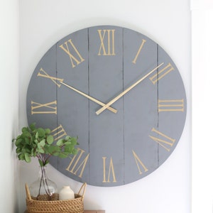 May include: A large gray wooden clock with Roman numeral markings in gold. The clock has gold-colored hands and is hanging on a white wall.