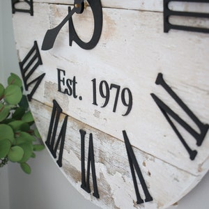 May include: White wooden clock with black Roman numeral hour markers and black hands. The clock face has a distressed look and includes the text "Est. 1979".