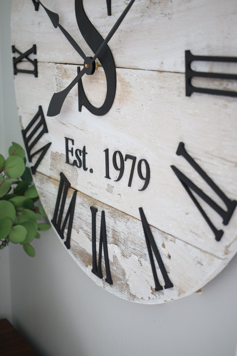 May include: White wooden clock with black Roman numeral hour markers and black hands. The clock face has a distressed look and includes the text "Est. 1979".