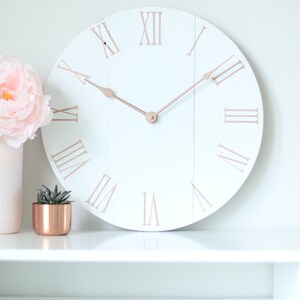 May include: White round clock with roman numerals in rose gold on a white shelf. The clock hands are also rose gold.