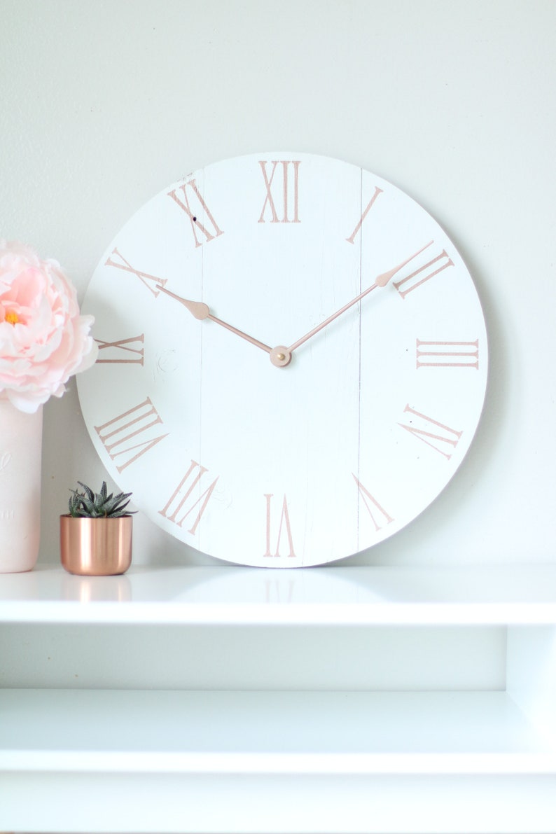 May include: White round clock with roman numerals in rose gold on a white shelf. The clock hands are also rose gold.