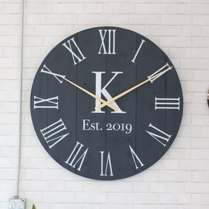 Può includere: Un grande orologio rotondo di colore grigio scuro con numeri romani bianchi e una lancetta delle ore e dei minuti dorata. Il quadrante dell'orologio presenta anche una lettera "K" bianca e il testo "Est. 2019".