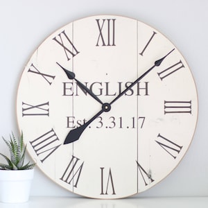 Può includere: Orologio bianco in stile farmhouse con indici orari in numeri romani neri e lancette nere. Il quadrante dell'orologio ha il testo "ENGLISH Est. 3.31.17".
