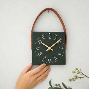 May include: A square dark green wall clock featuring a white circular numeral track with numbers one through twelve and thin gold-colored hands. The clock is suspended by a brown leather strap attached to the top sides of the square frame.