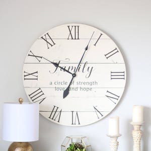 May include: White farmhouse style clock with black Roman numeral hour markers and black hands. The clock face has a wood plank design and the text "Family, a circle of strength love and hope".