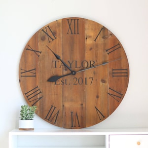 May include: A large, rustic wooden clock with Roman numeral markings and black hands. The clock face is personalized with the name "TAYLOR" and the date "Est. 2017".