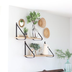 May include: Two wooden shelves with black leather straps, mounted on a white wall. The shelves are decorated with potted plants, a vase with greenery, a wicker basket, and a small round mirror.