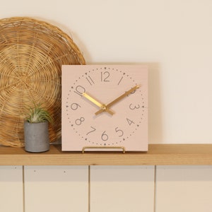 May include: A pink square clock with gold hands and black numbers on a wooden shelf. The clock is sitting on a gold metal stand.