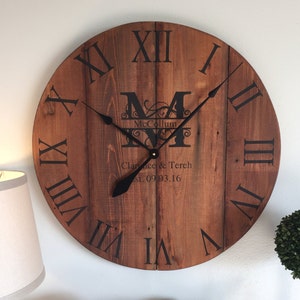 May include: A large, rustic wooden clock with Roman numeral markings. The clock face is brown with a black stain and features a personalized monogram with the initials "M" and "M" and the name "McCollum". The clock also includes the text "Clarence & Tereh Est. 09.03.16".