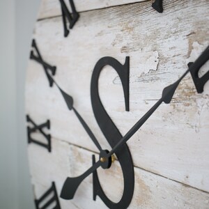 May include: A whitewashed wooden clock face with black Roman numeral hour markers and black hands. The clock face also features a large black letter "S" and the text "Est".