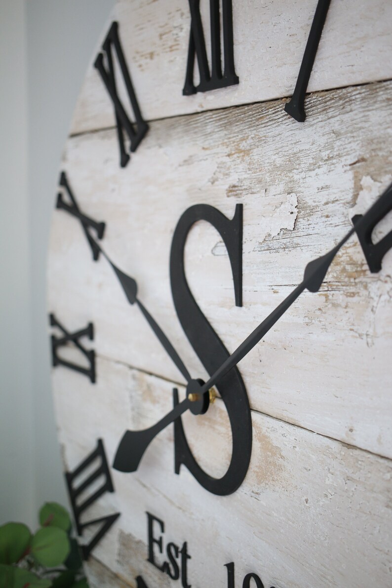 May include: A whitewashed wooden clock face with black Roman numeral hour markers and black hands. The clock face also features a large black letter "S" and the text "Est".