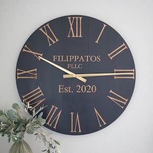 May include: A large, round, black wooden clock with gold Roman numeral hour markers and gold hands. The clock face also includes the text "Filippatos PLLC Est. 2020".