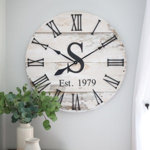 May include: A whitewashed wooden clock with Roman numerals and black hands. The clock has a large black letter "S" in the center and the text "Est. 1979" below it.
