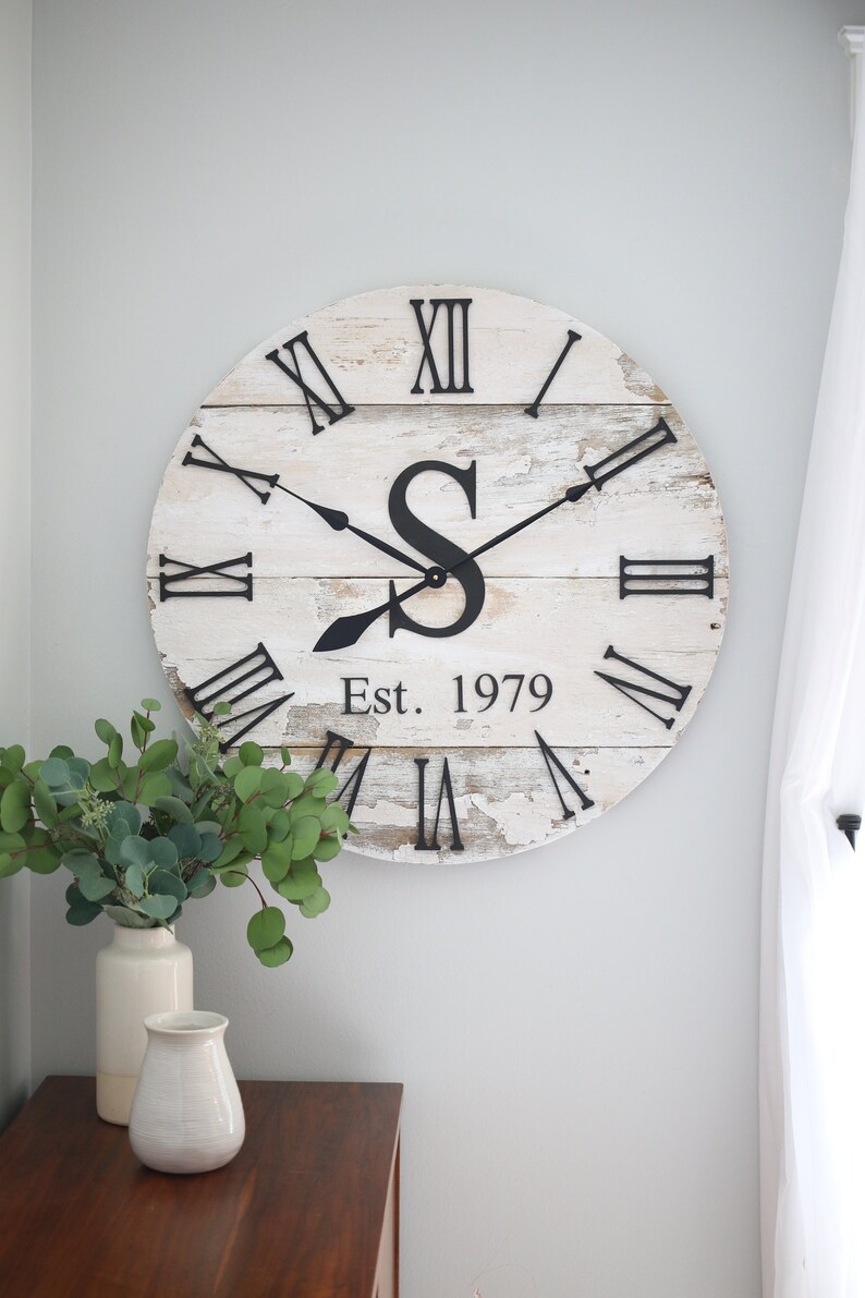 May include: A whitewashed wooden clock with Roman numerals and black hands. The clock has a large black letter "S" in the center and the text "Est. 1979" below it.