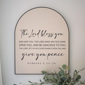 May include: A beige arch-shaped wooden sign with black text that reads "The Lord bless you and keep you; The Lord make his face shine upon you, and be gracious to you; The Lord lift up his countenance upon you and give you peace. Numbers 6:24-26"