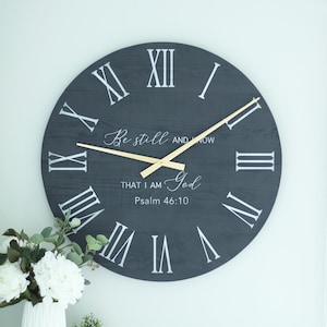 May include: A dark grey wooden clock with Roman numeral markings and gold hands. The clock face features the text "Be still and know that I am God, Psalm 46:10".