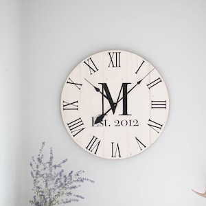May include: White round clock with black Roman numeral markings and black hands. The clock face also features a black letter "M" and the text "Est. 2012".