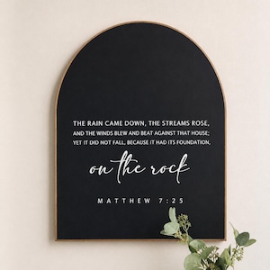 May include: Black arched wooden sign with the text "THE RAIN CAME DOWN...on the rock MATTHEW 7:25" in white. The sign has a dark background and a light brown wooden frame. A sprig of greenery is in the lower right corner.