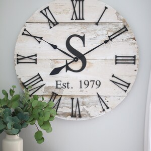May include: A white, distressed wood clock with black Roman numeral hour markers and black hands. The clock face also features a black letter "S" and the text "Est. 1979".
