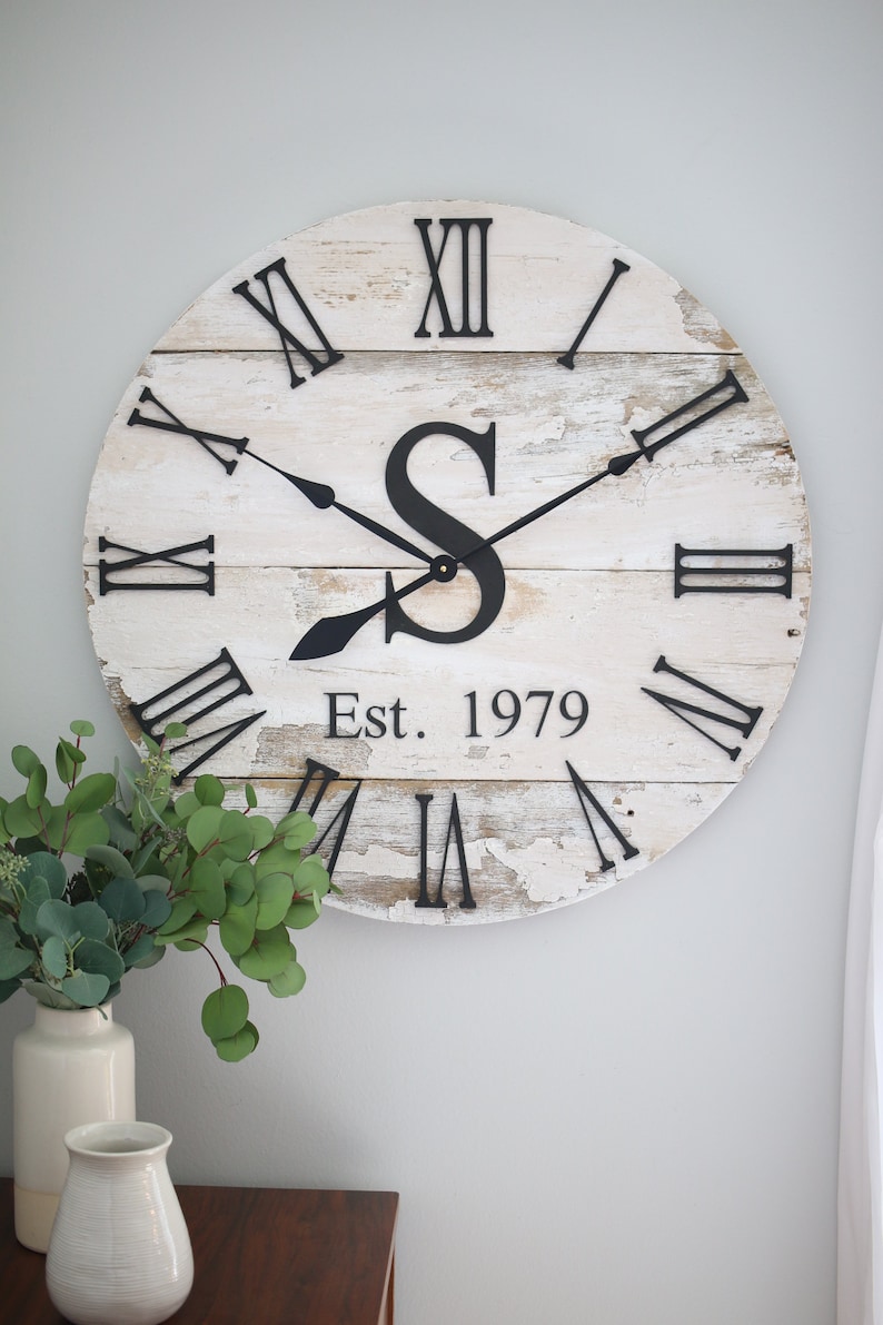 May include: A white, distressed wood clock with black Roman numeral hour markers and black hands. The clock face also features a black letter "S" and the text "Est. 1979".