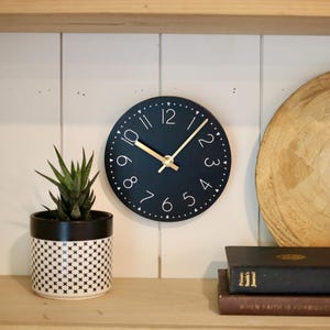 May include: A black round clock with white numbers and gold hands. The clock is hanging on a white wall shelf with a potted plant and books.