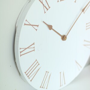 May include: White round clock face with Roman numeral markings in a copper color. The clock hands are also copper colored.
