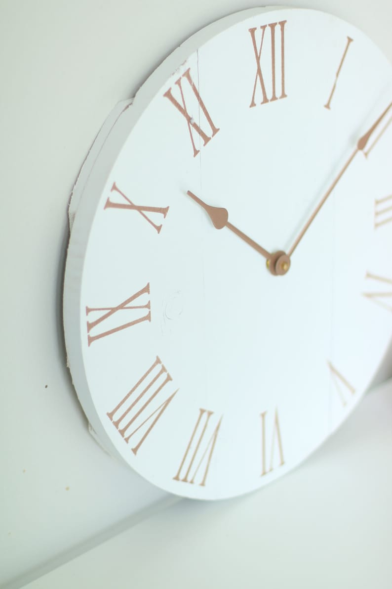 May include: White round clock face with Roman numeral markings in a copper color. The clock hands are also copper colored.