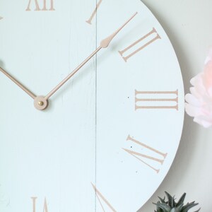 May include: A light blue round clock with roman numeral markings in a copper color. The clock hands are also copper colored. The clock is on a white surface with a small copper colored pot with a green succulent plant.