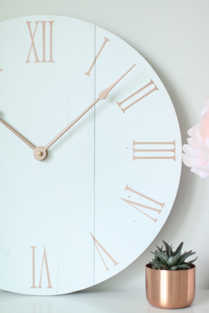 May include: A light blue round clock with roman numeral markings in a copper color. The clock hands are also copper colored. The clock is on a white surface with a small copper colored pot with a green succulent plant.