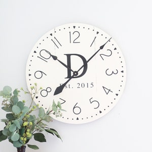 May include: White farmhouse style clock with black Roman numerals and hands. The clock face features a black letter "D" and the text "Est. 2015".