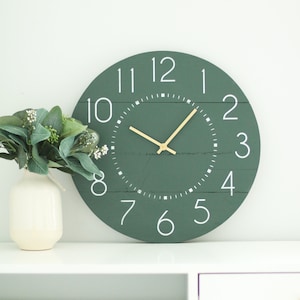 May include: A green round clock with white numbers and a gold-colored hour and minute hand. The clock is on a white surface.