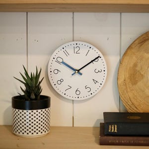 May include: A white round clock with black numbers and hands. The clock is hanging on a white wall with a wooden shelf above it.