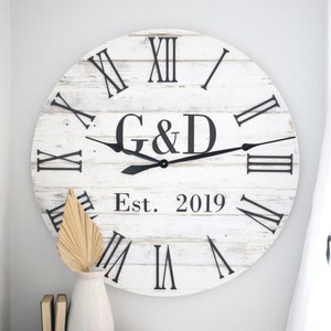 May include: White wooden clock with black Roman numeral markings and black hands. The clock face also includes the initials "G&D" and the text "Est. 2019".