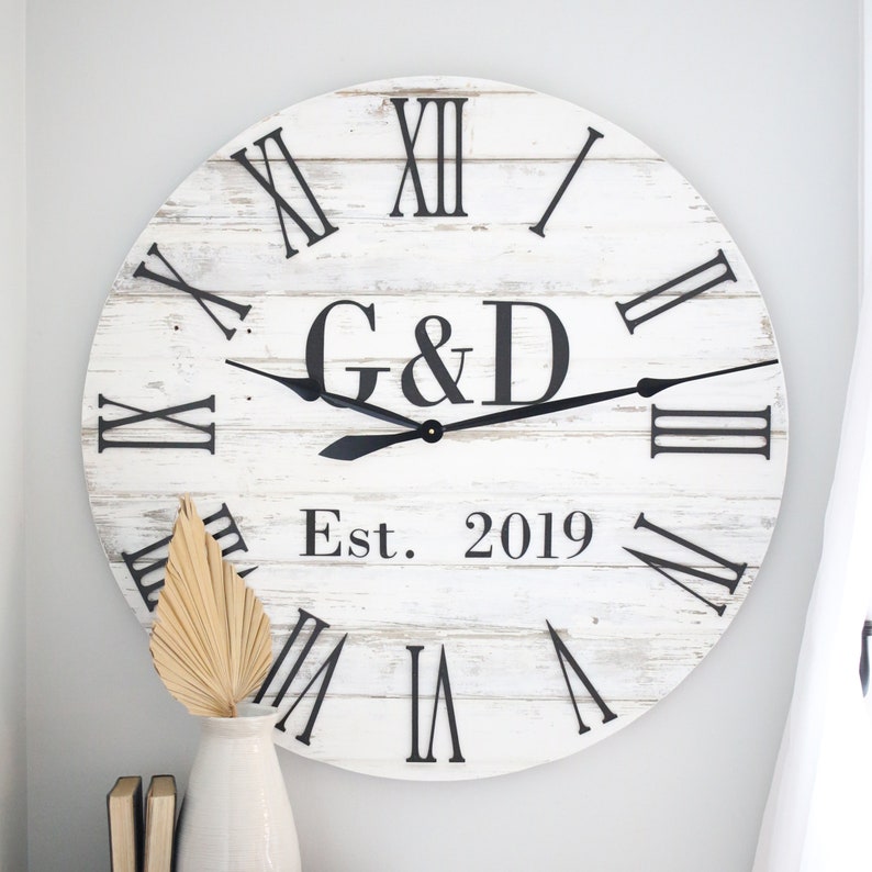 May include: White wooden clock with black Roman numeral markings and black hands. The clock face also includes the initials "G&D" and the text "Est. 2019".