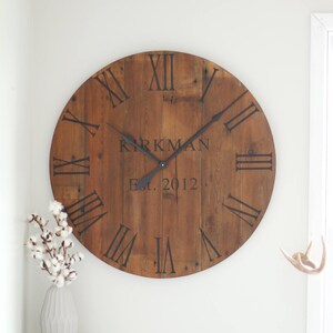 May include: A large, round, wooden clock with Roman numeral markings and black hands. The clock face is made of reclaimed wood and has a personalised inscription that reads "Kirkman Est. 2012".