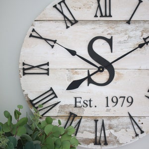 May include: A white wooden clock with black Roman numeral markings and black hands. The clock has a distressed finish and features a black letter "S" and the text "Est. 1979".