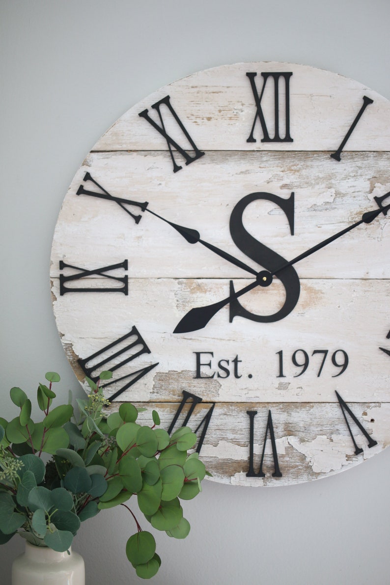 May include: A white wooden clock with black Roman numeral markings and black hands. The clock has a distressed finish and features a black letter "S" and the text "Est. 1979".