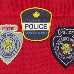 May include: Three embroidered patches, one blue with "POLICE" and "BROKEN ARROW OKLAHOMA" in white, one blue with "POLICE" and "Canadian Pacific" in white, and one gray with "FIRE" and "BROKEN ARROW" in yellow.