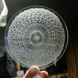 May include: A clear, circular glass object with an intricate pattern of small, white, crescent-shaped designs. The design radiates from a central point, creating a mesmerizing visual effect. The object is held up by a hand.