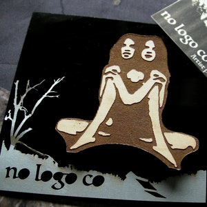 May include: A laser-cut wooden pin featuring a stylized figure with a large head and two eyes. The figure is sitting with its legs crossed and its hands clasped in front of it. The pin is on a black background with the text "no logo co" printed in white.