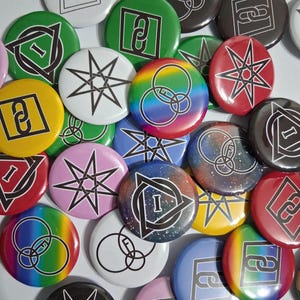 Puede incluir: Una colección de botones redondos y coloridos con varios diseños. Algunos botones presentan formas geométricas, estrellas y la letra "I" dentro de un triángulo. Otros muestran patrones de arcoíris, galaxias y abstractos. Los botones son en tonos verdes, rojos, azules, amarillos y rosas.