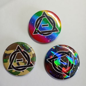 Puede incluir: Tres botones redondos con un diseño de triángulo y círculo en blanco y negro. Los botones tienen diferentes fondos: uno es arcoíris, otro es camuflaje y otro es un estampado de galaxia.