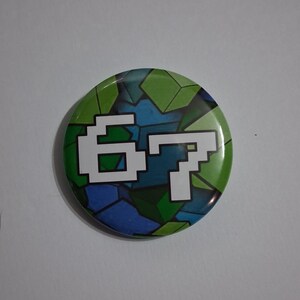 May include: A circular badge with a pixelated design. The background is a mosaic of green, blue, and dark green geometric shapes. The number "67" is displayed in white, pixelated font, centered on the badge.
