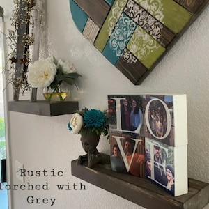May include: Rustic wooden shelves with a torched grey finish. The shelves are decorated with a white heart-shaped wall art, a small potted plant, a white vase with white flowers, and a wooden block sculpture that spells "LOVE" with photos of people in each letter.