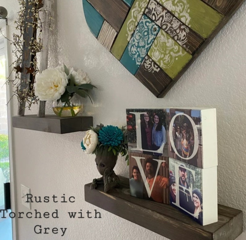 May include: Rustic wooden shelves with a torched grey finish. The shelves are decorated with a white heart-shaped wall art, a small potted plant, a white vase with white flowers, and a wooden block sculpture that spells "LOVE" with photos of people in each letter.