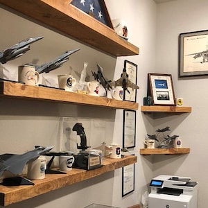 May include: A collection of model airplanes and mugs displayed on wooden shelves. The shelves are mounted on a white wall. The airplanes are various models of fighter jets. The mugs are decorated with aviation themes. The shelves are made of unfinished wood and have a rustic look.