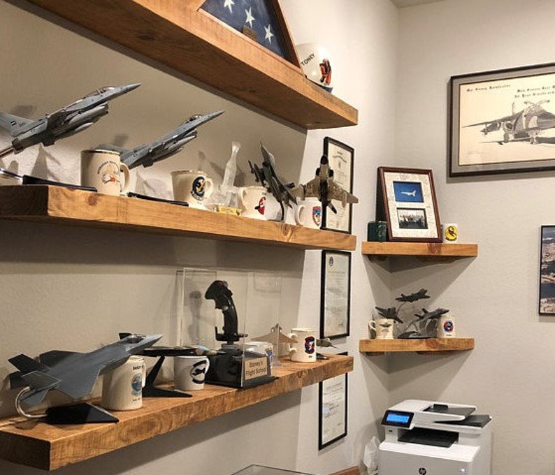 May include: A collection of model airplanes and mugs displayed on wooden shelves. The shelves are mounted on a white wall. The airplanes are various models of fighter jets. The mugs are decorated with aviation themes. The shelves are made of unfinished wood and have a rustic look.