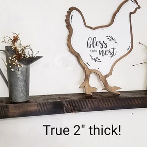 May include: A dark brown wooden shelf with a white chicken silhouette sign that says "bless this nest". The shelf is 2 inches thick.