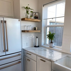 May include: White kitchen with a stainless steel refrigerator, white cabinets, and a white quartz countertop. There are two wooden shelves on the wall above the countertop, with a plant, glasses, and a small decorative object on the shelves. A window with a white blind is on the right side of the image, with a plant in a white pot on the windowsill.