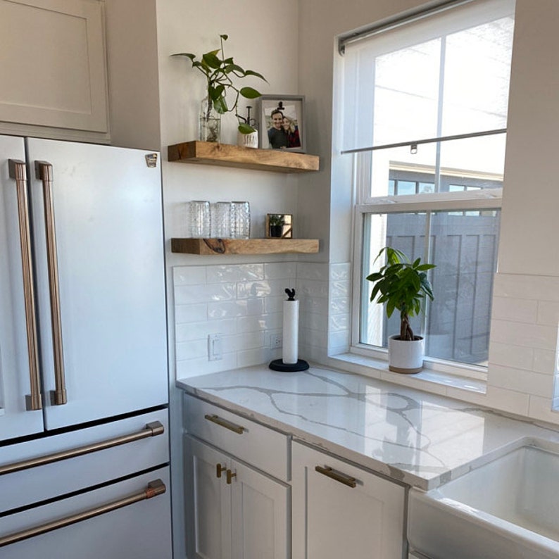 May include: White kitchen with a stainless steel refrigerator, white cabinets, and a white quartz countertop. There are two wooden shelves on the wall above the countertop, with a plant, glasses, and a small decorative object on the shelves. A window with a white blind is on the right side of the image, with a plant in a white pot on the windowsill.